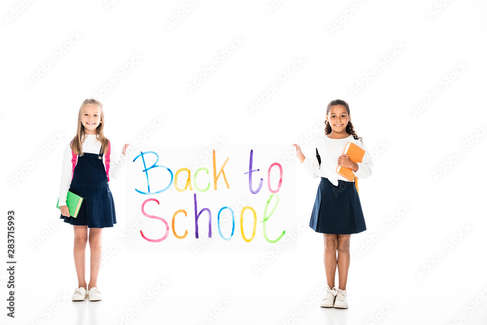 full length view of two multicultural schoolgirls holding books and placard with back to school inscription on white background full length view of two multicultural schoolgirls holding books and placard with back to school inscription on white background