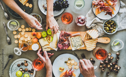 Mid-summer picnic with wine and snacks. Flat-lay of charcuterie and cheese board, rose wine, nuts, olives and peoples hands over concrete table background, top view. Family, friends holiday gathering photo