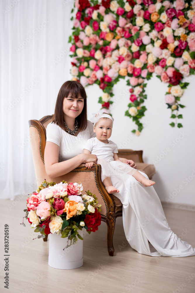 mother's day concept - happy beautiful mother and her cute little daughter sitting on vintage sofa over flowers wall mother's day concept - happy beautiful mother and her cute little daughter sitting on vintage sofa over flowers wall