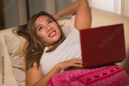 young beautiful and natural Asian Indonesian woman in pajamas pants relaxed on couch networking using laptop computer shopping online having fun in dreamy face expression