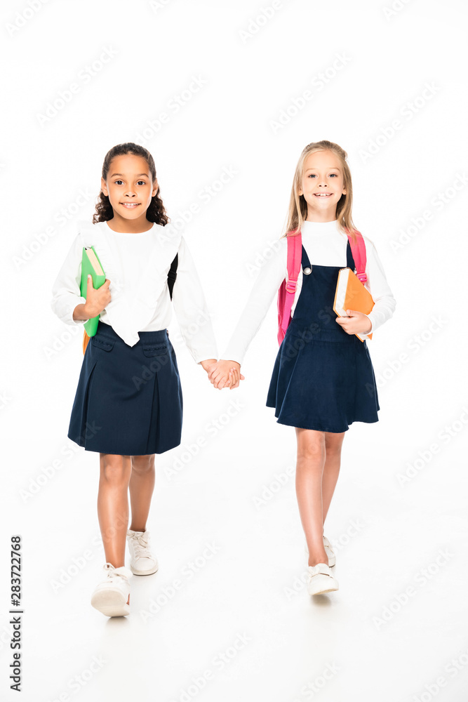 full length view of two smiling multicultural schoolgirls walking while holding hands on white background full length view of two smiling multicultural schoolgirls walking while holding hands on white background