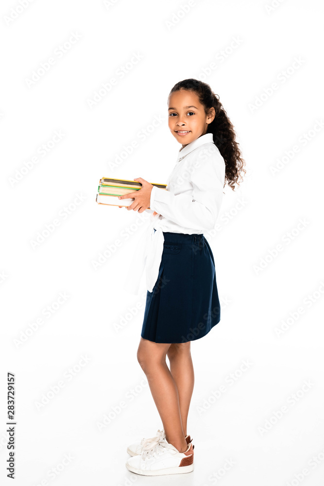 full length view of adorable african american schoolgirl holding books and smiling at camera on white background full length view of adorable african american schoolgirl holding books and smiling at camera on white background