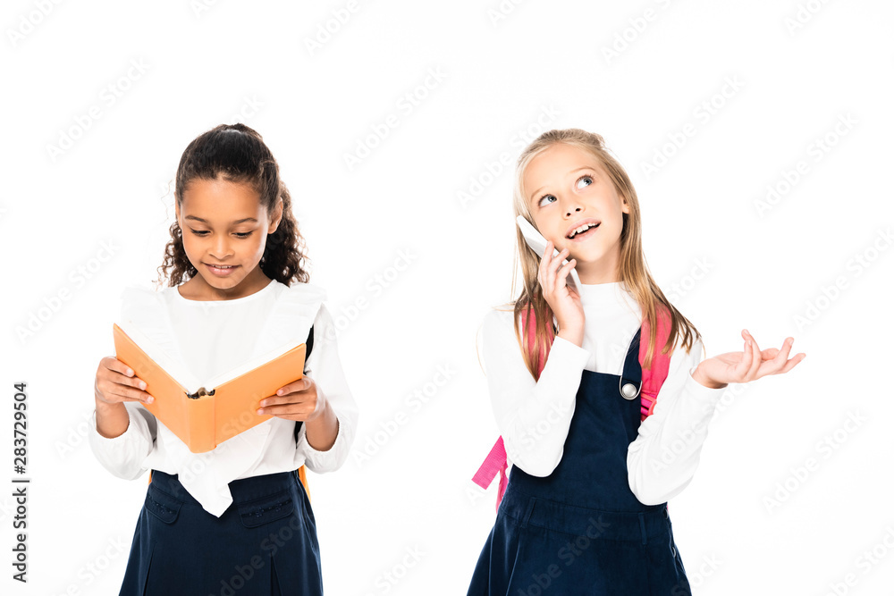 cheerful schoolgirl talking on smartphone near african american friend reading book isolated on white cheerful schoolgirl talking on smartphone near african american friend reading book isolated on white