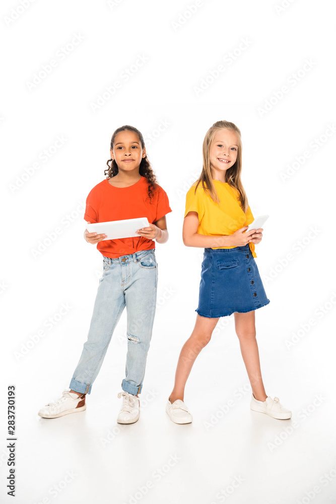 full length view of two adorable multicultural friends holding smartphone and digital tablet on white background
