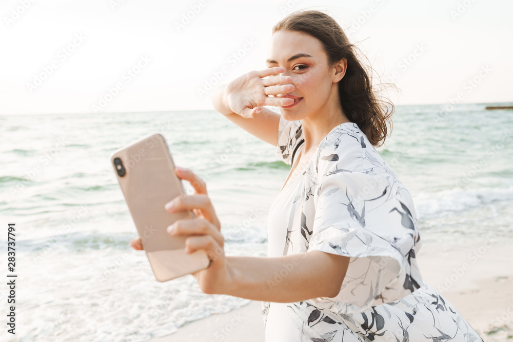 Pleased happy young beautiful woman at the beach walking in a beautiful sunny morning take a selfie by mobile phone.