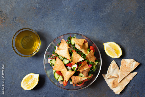 Traditional Fattoush salad with vegetables and pita bread. Levantine, Arabic, Middle Eastern cuisine. Served in a glass plate with lemon, pita and olive oil. Dark background. Top view. Space for text