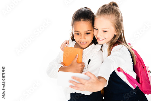 cheerful schoolgirl hugging african american friend and taking selfie isolated on white
