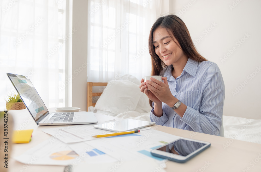 Asian businesswoman drinking coffee and while working with laptop on table in bedroom at home.Work at home concept.work from home concept Asian businesswoman drinking coffee and while working with laptop on table in bedroom at home.Work at home concept.work from home concept