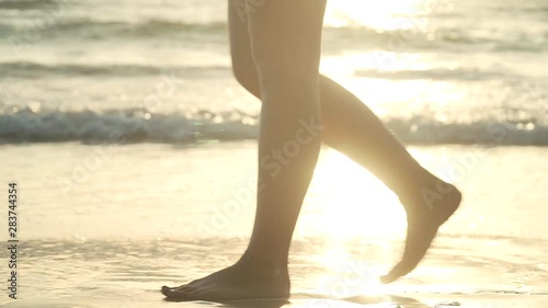Close-up of legs of young woman walking along tropical beach at sunset. Foamy sea waves gentrly wash sand and feet, warm soft evening sun reflects on wet surface. Slow motion footage.