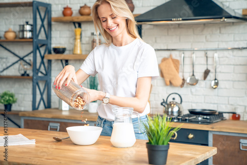 Pretty young girl in a kitchen prepares a healthy breakfast with corn flakes and fresh milk. Woman has a breakfast on stylish cozy kitchen at the morning. Diet concept.