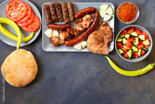 Traditional Serbian and Balkan grilled meat called mesano meso. Balkan barbeque (rostilj) served with Serbian salad, hot peppers, bread, tomato, onions, and paprika powder. Dark background. Top view photo