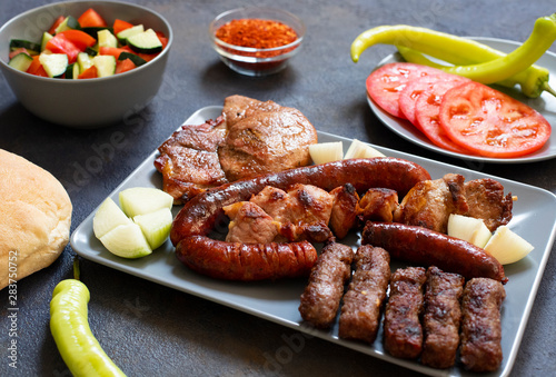 Traditional Serbian and Balkan grilled meat called mesano meso. Balkan barbeque (rostilj) served with Serbian salad, hot peppers, bread, tomato, onions, and paprika powder. Dark background. Close-up photo