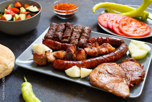Traditional Serbian and Balkan grilled meat called mesano meso. Balkan barbeque (rostilj) served with Serbian salad, hot peppers, bread, tomato, onions, and paprika powder. Dark background. Close-up photo