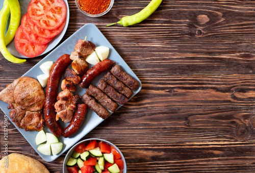 Traditional Serbian and Balkan grilled meat called mesano meso. Balkan barbeque (rostilj) served with Serbian salad, hot peppers, bread, tomato, onions, and paprika powder. Wooden background. Top view photo