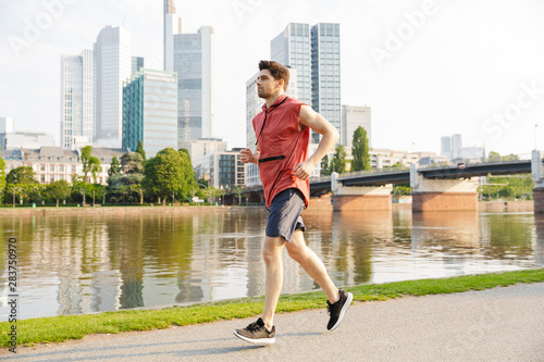 Photo of young serious man running while working out near city riverfront
