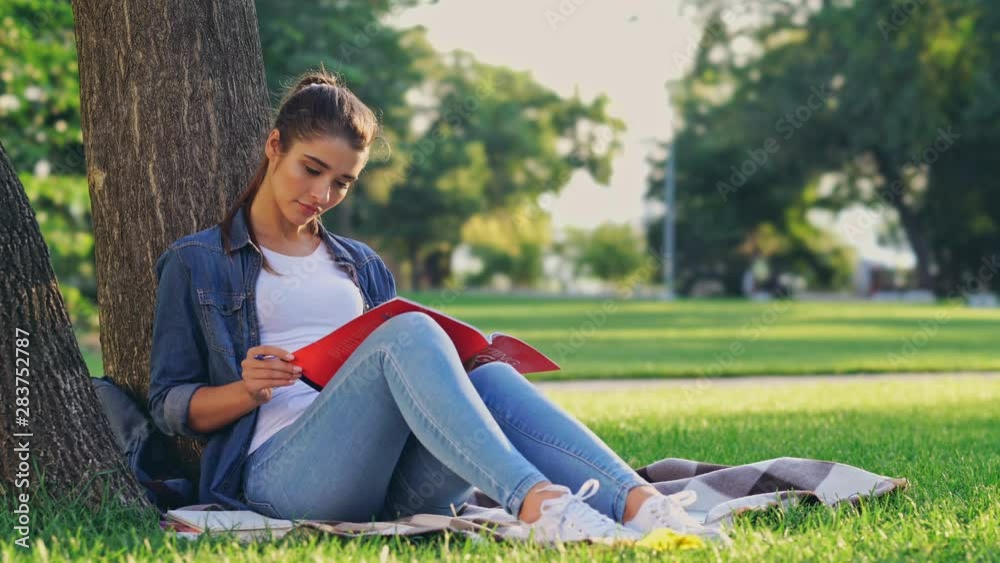 Smiling brunette woman reading magazine and writing something while sitting near the tree on grass in park