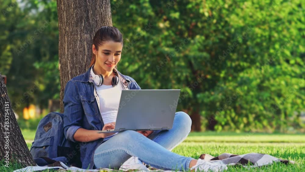 Pleased brunette woman using laptop computer while sitting near the tree on grass in park Pleased brunette woman using laptop computer while sitting near the tree on grass in park