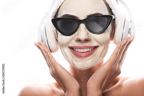 front view of nude woman in cosmetic hair band and sunglasses with facial mask listening music in headphones isolated on white