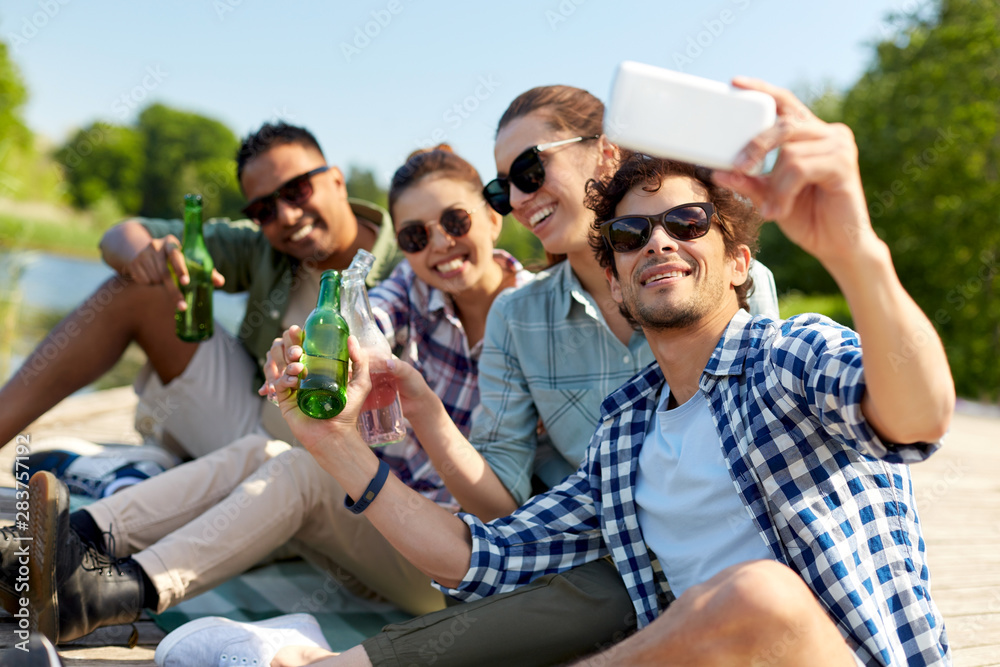 Obraz premium leisure, picnic and technology concept - friends with drinks taking selfie by smartphone on lake pier in summer park