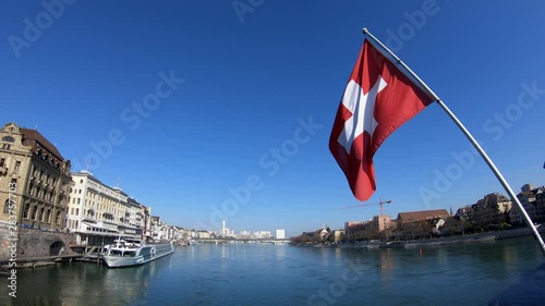 Basel, Switzerland - March 22, 2018: 4K. Swiss flag waving in the wind on the Käppelijoch bridge, which crosses the Rhein river. Buildings around the river, people walking. Switzerland.
