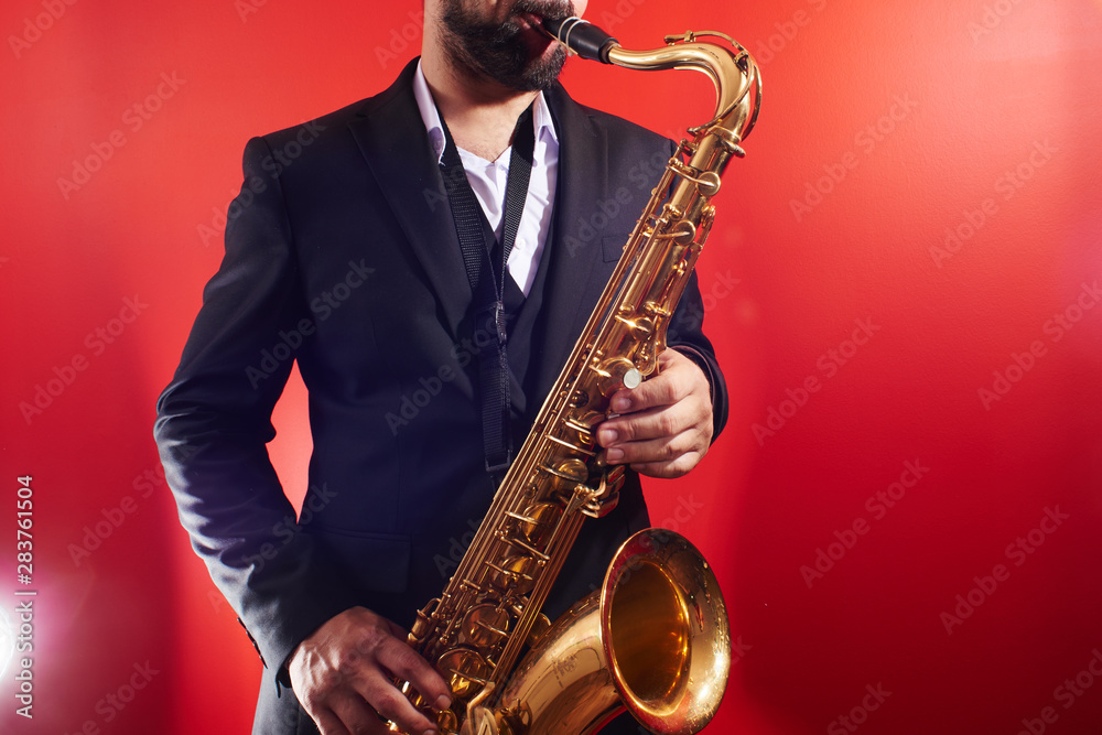 Fototapeta premium Portrait of professional musician saxophonist man in suit plays jazz music on saxophone, red background in a photo studio
