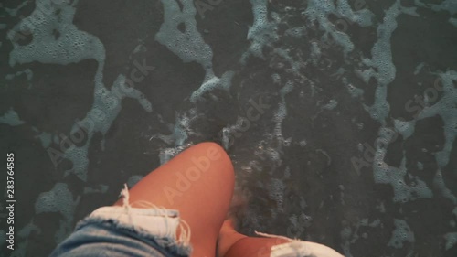Top view close-up of young woman legs standing on sea shore at sunset. Foamy sea waves gentrly wash sand and feet, warm soft evening sun reflects on wet surface. First person view slow motion footage.