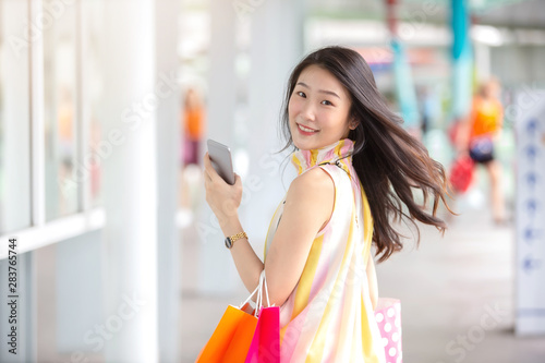 Young and beautiful Asian woman with colourful shopping bag holding her mobile phone while shopping in the shopping mall.
