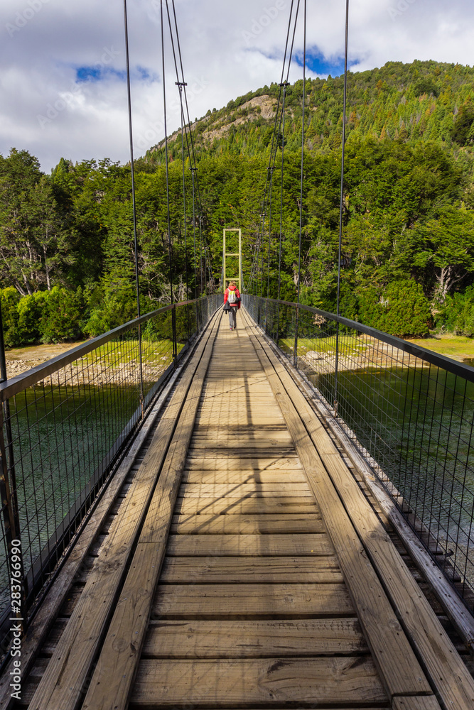Rear view of a young woman practicing hiking wearing a backpack crossing a wooden suspension bridge over a wide river in Los Alerces National Park, Patagonia, Argentina