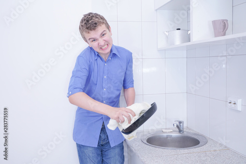 Fotografie A cute teenager boy washing dishes at white kitchen alone with sad bad unhappy f