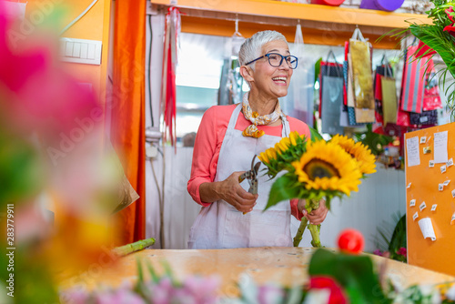 Smiling portrait of an attractive florist business woman owner at a flower shop market working and making a new floral arrangement during a sunny day. Small business owner.