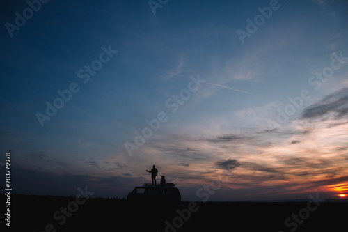 Silhouette of a man with a guitar and a woman in a hat stand on the roof of a car on the background of the sunset. Silhouette romance concept.