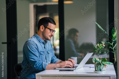 Serious pensive thoughtful focused young casual businessman or entrepreneur in office looking at and working with laptop making and typing serious important business email