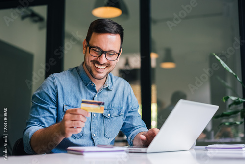 Happy cheerful smiling young adult man doing online shopping or e-shopping satisfied entrepreneur making online payment paying for service or goods self employed freelancer collecting fee photo