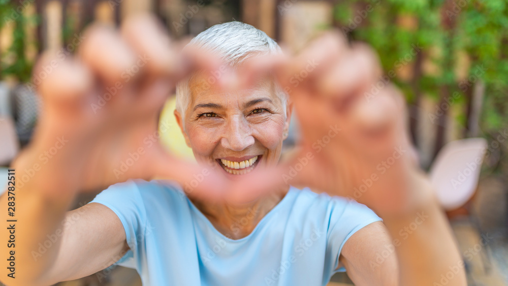 Cute senior old woman making a heart shape with her hands and fingers. Perfect, nice, aged, old, pretty woman, lover in t-shirt making, showing heart figure with fingers, looking at camera - obrazy, fototapety, plakaty 