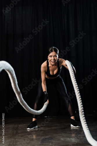 selective focus of sportswoman working out with battle ropes on black