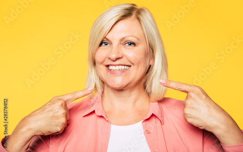 Perfect result of dental surgery. Beautiful happy blonde senior woman is pointing on her toothy healthy smile and looks on camera
