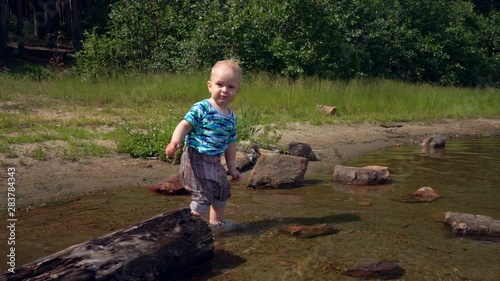 Little boy steps on the water of a forest lake on a sunny summer day. Child plays with stones that he is trying to get from the bottom of the lake from clear water