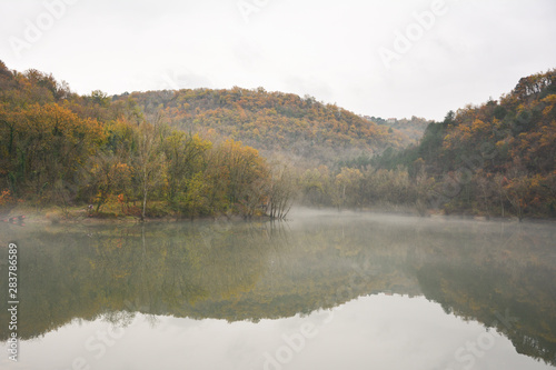 Moody lake in the fog, surrounded by colorful forests. Wood in autumn. Lake in Europe, Slovenia, Koper, Vanganel. Vanganelsko jezero.