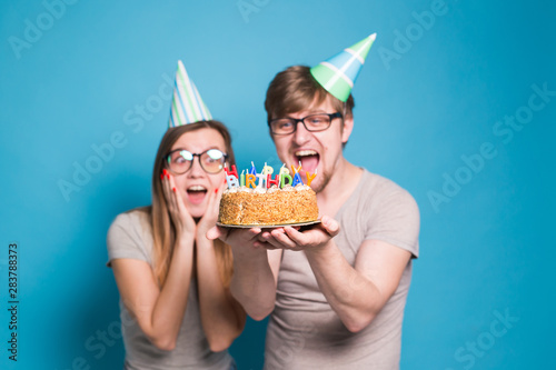 Funny nerd man and woman are wearing holidays caps and glasses holding birthday cake with candles over blue background