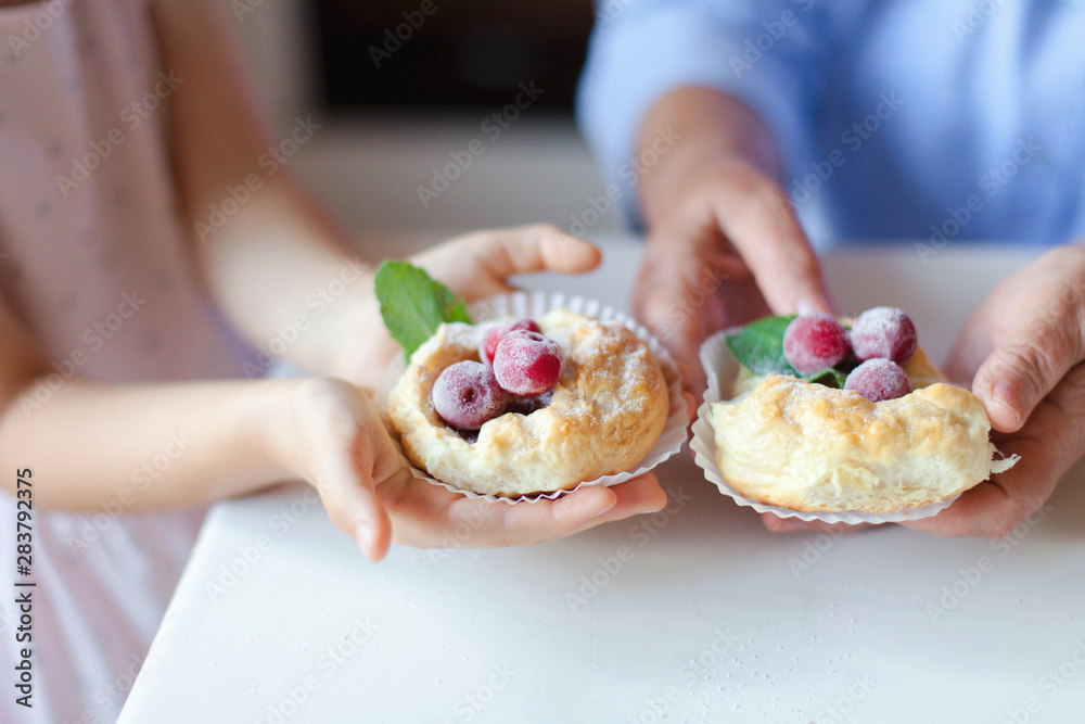 Senior woman and child girl with homemade pastries at kitchen. Baked buns with berries and mint leaves. Sweets with powdered sugar. Hands of kid and retired grandmother.