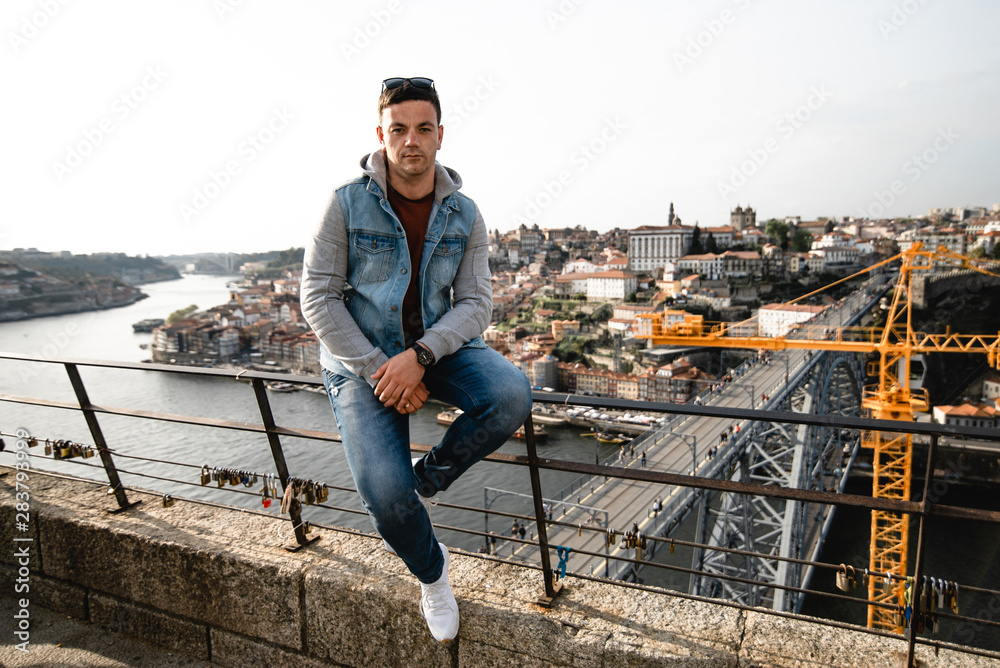 Handsome man staying on the bridge on the city background. Tourist is walking on the quay. The young guy admires the beauty of the old city in Portugal. Handsome man staying on the bridge on the city background. Tourist is walking on the quay. The young guy admires the beauty of the old city in Portugal.