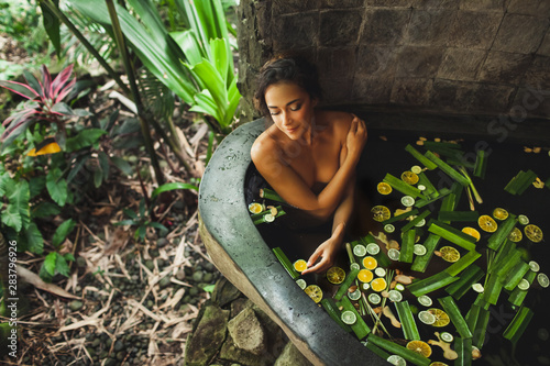 Beautiful young woman enjoying in outdoor spa. Luxury stone bath tub with jungle view. Natural organic tropical ingredients in the water: ginger, lime, orange and sea salt. Beauty treatment concept.