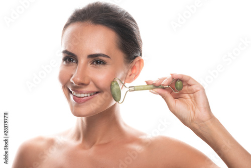 naked young woman using facial jade roller and smiling isolated on white