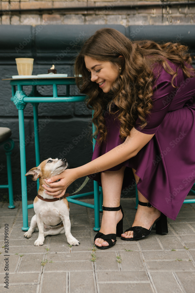 Dog and girl in street cafe. Beautiful woman in purple dress pettin small cute dog sitting under the table. Street cafe