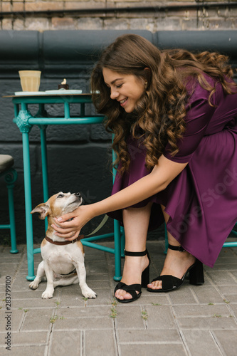 Dog and girl in street cafe. Beautiful woman in purple dress pettin small cute dog sitting under the table. Street cafe
