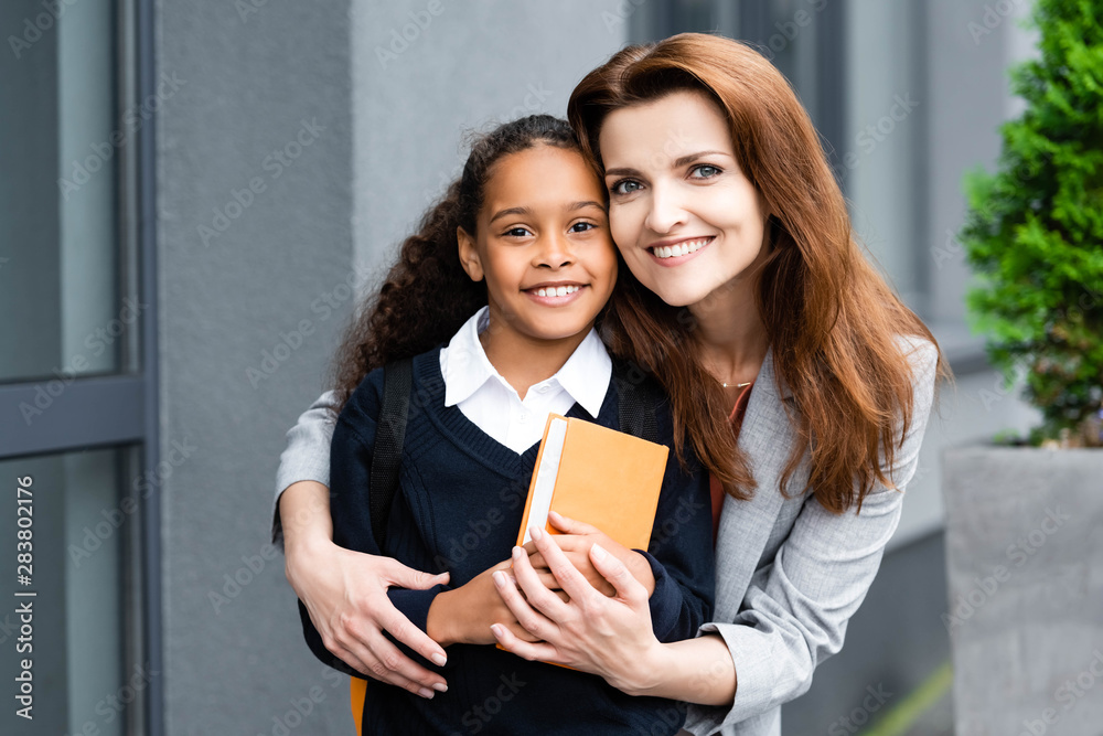 happy mother hugging adopted african american daughter while standing near scholl together