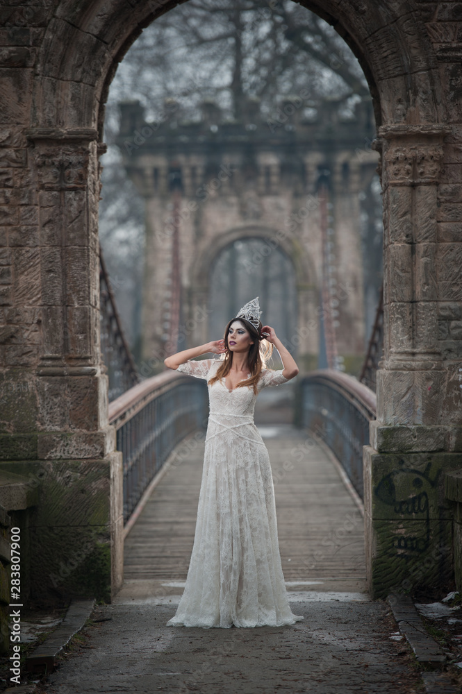 Obraz premium Lovely young lady wearing elegant white dress enjoying the beams of celestial light and snowflakes falling on her face. Pretty brunette girl in long wedding dress posing on a bridge in winter scenery