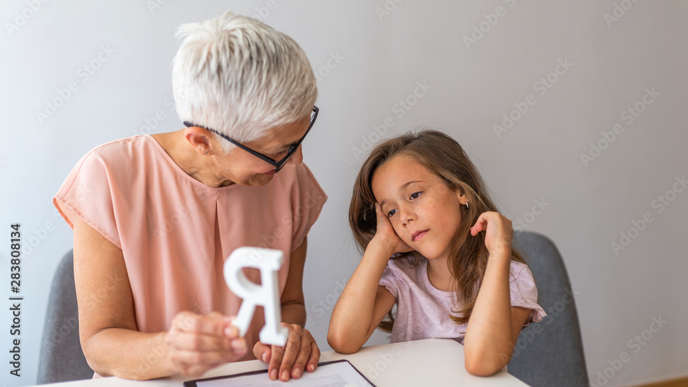 Little girl with teacher on private lesson. A child with development problems with a professional speech therapist during a meeting. Tutor holding a letter 'R'. Little girl with teacher on private lesson. A child with development problems with a professional speech therapist during a meeting. Tutor holding a letter 'R'.