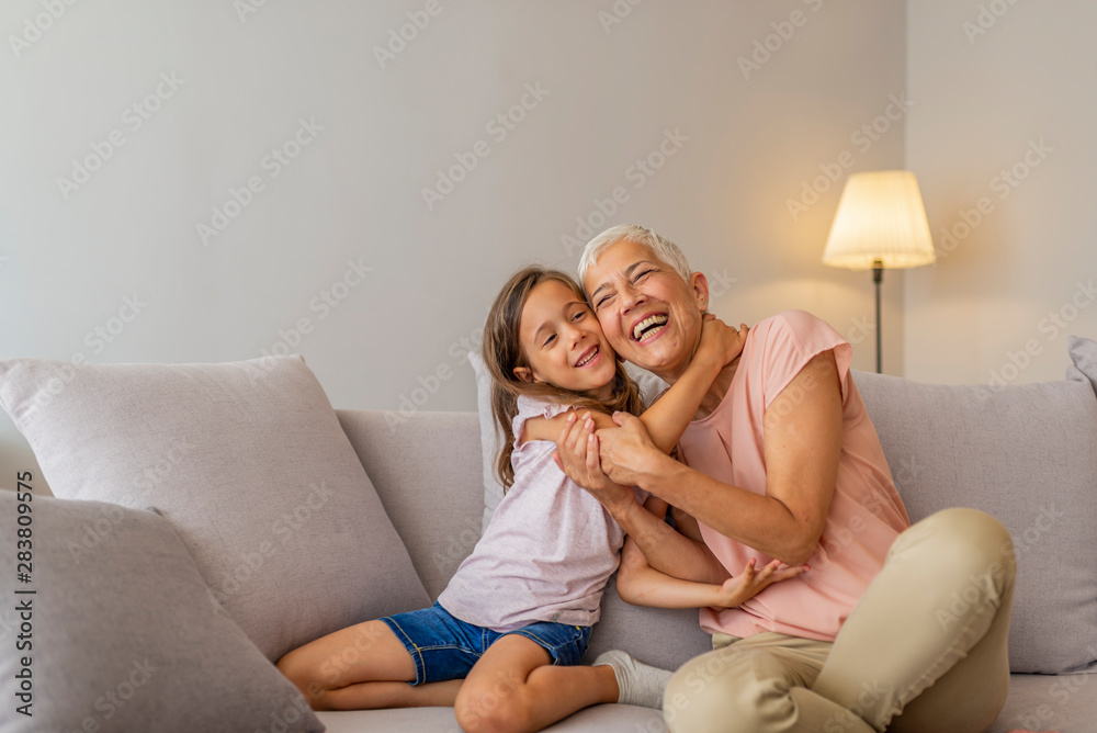 Grandmother and granddaughter hug. Little cute girl and her grandmother are spending time together at home. Having fun, hugging and smiling while sitting on sofa. - obrazy, fototapety, plakaty 