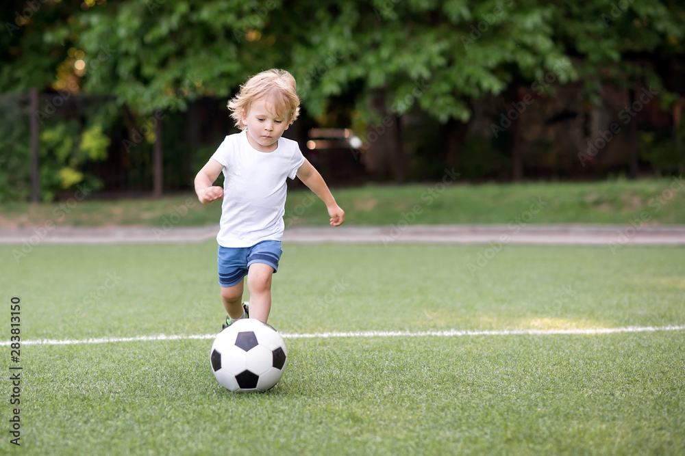 Little football player: blonde child in white shirt and blue shorts running along the green soccer field ready to kick ball. Young sportsman and active childhood concept